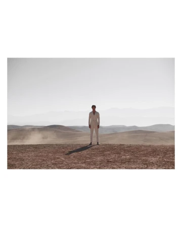Full body shot of an adult male in white suit standing on arid landscape with distant mountains, under bright daylight.