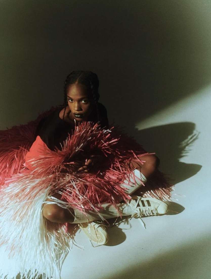 Young woman poses in black top and Adidas sneakers surrounded by a fuzzy pink and white fringed garment on a light grey backdrop.