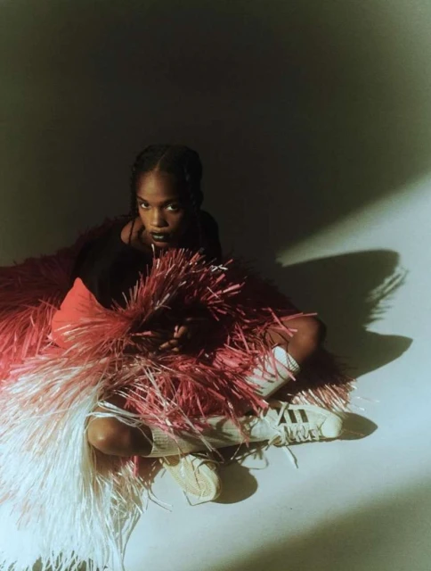 Young woman poses in black top and Adidas sneakers surrounded by a fuzzy pink and white fringed garment on a light grey backdrop.