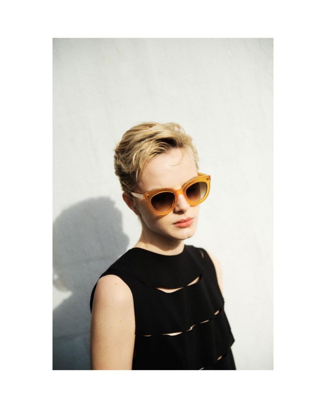Woman with short blonde hair poses wearing sunglasses and a black, sleeveless top against a white wall in a medium studio shot.