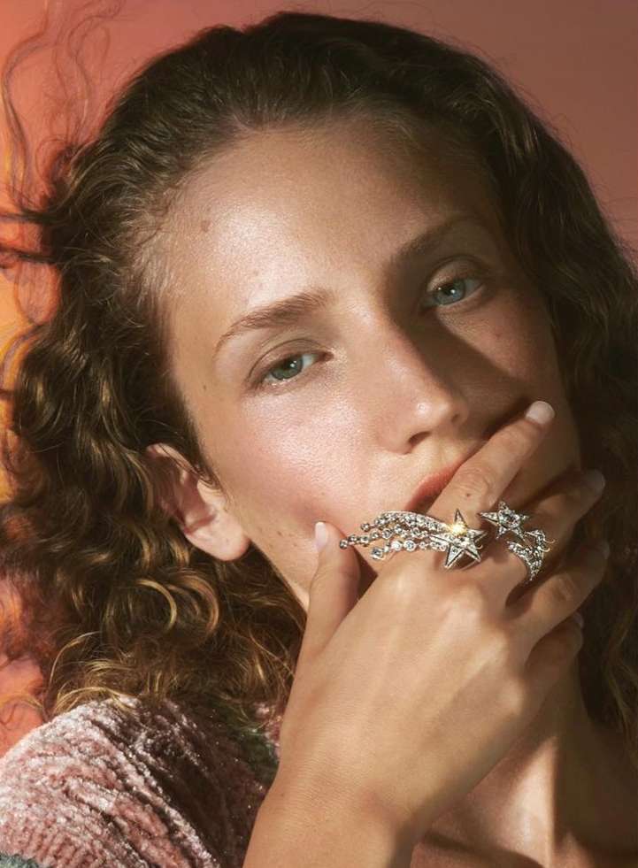 Woman with curly hair and sparkly star rings poses indoors with coral backdrop in close-up studio portrait for beauty or fashion concept.