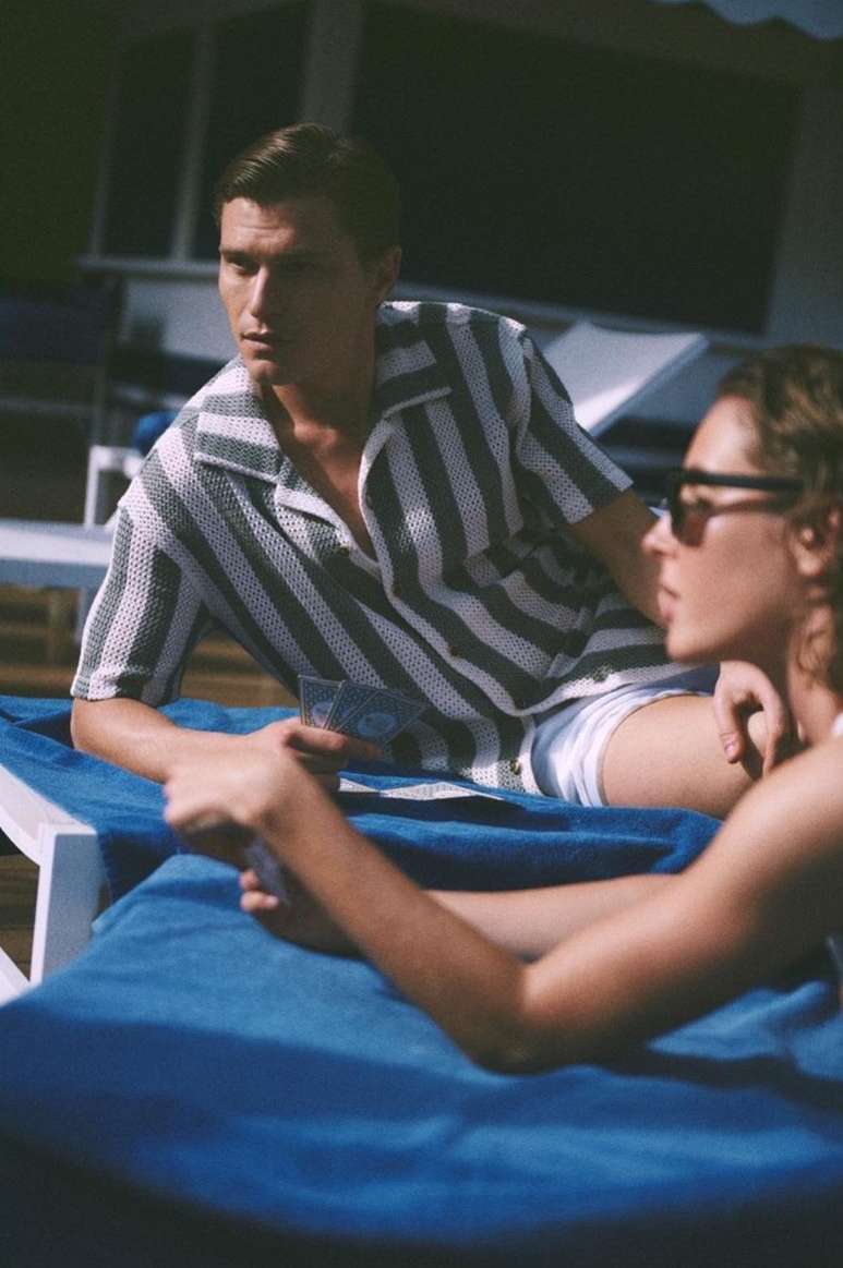 Two people relax on blue lounge chairs outdoors. One man wears a striped shirt, a woman wears sunglasses; blue loungers.