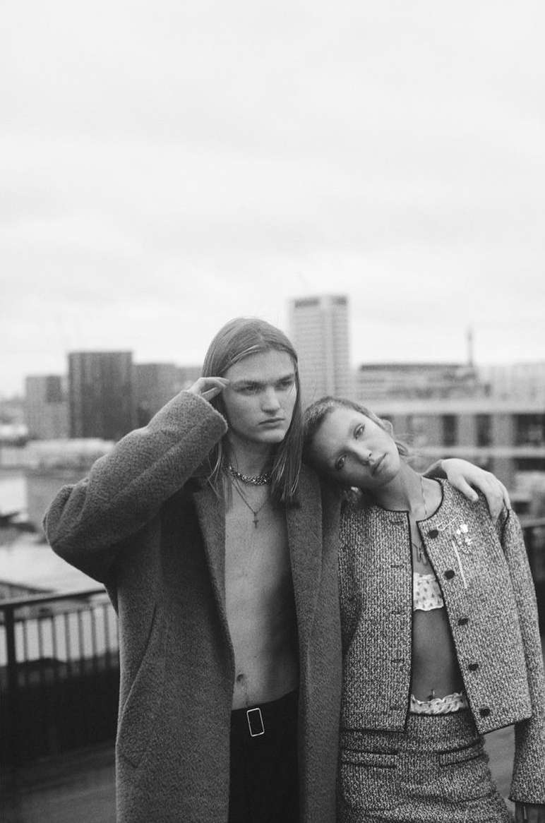 Black and white fashion portrait of a young man and woman posing in stylish clothing on a rooftop in front of a city skyline.