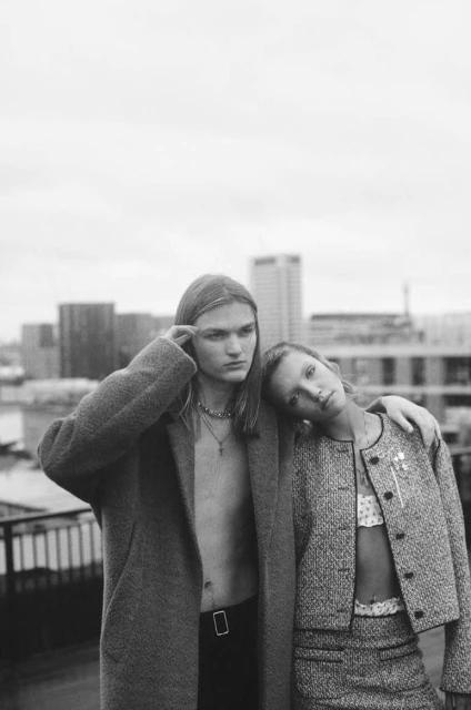Black and white fashion portrait of a young man and woman posing in stylish clothing on a rooftop in front of a city skyline.