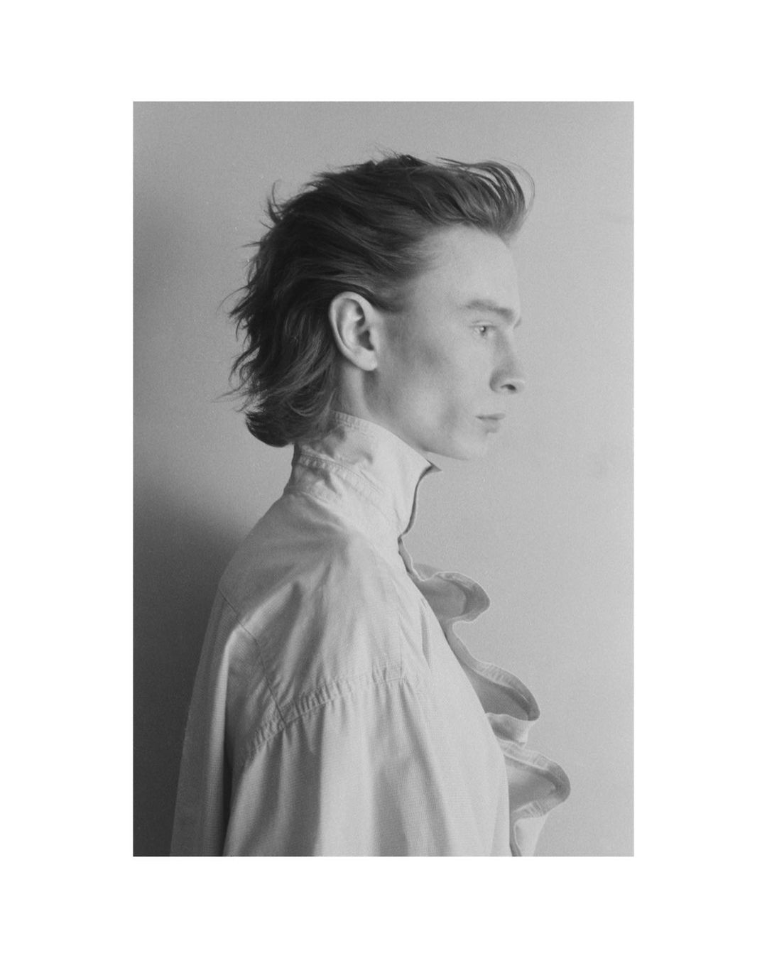 Monochrome portrait of a young adult male in profile, featuring ruffled high-collar shirt, shoulder-length hair, and neutral facial expression.