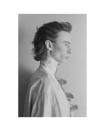 Monochrome portrait of a young adult male in profile, featuring ruffled high-collar shirt, shoulder-length hair, and neutral facial expression.