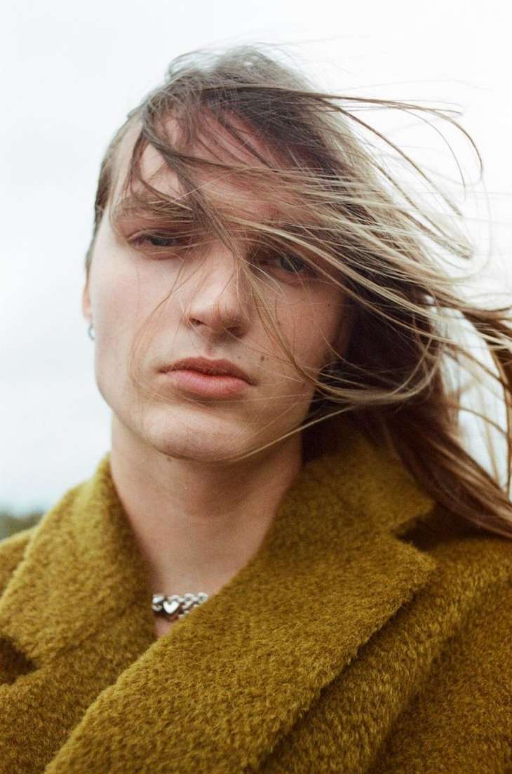 Portrait of a young man with long blonde hair blowing across his face wearing an olive green textured coat by Olivier Yoan