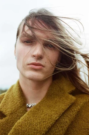 Portrait of a young man with long blonde hair blowing across his face wearing an olive green textured coat by Olivier Yoan