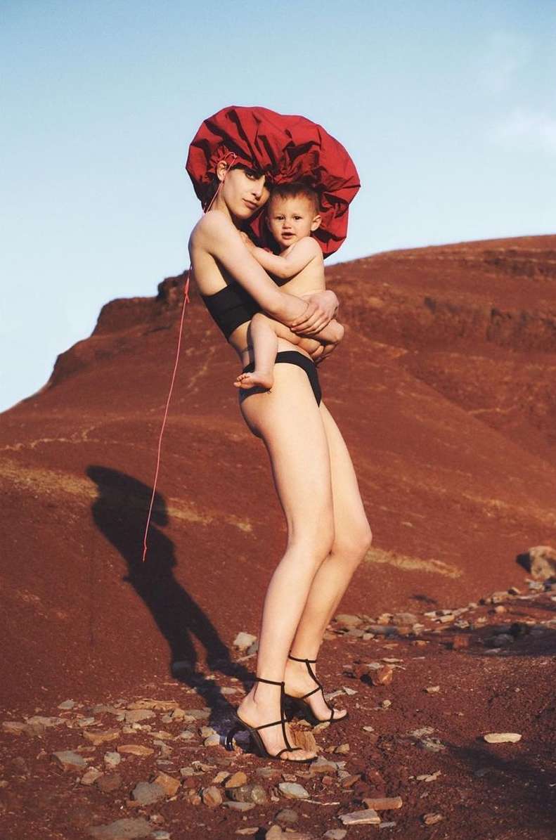 A woman in black swimwear and a large red bonnet holds a baby in the desert landscape against a light blue sky in a fashion shot.