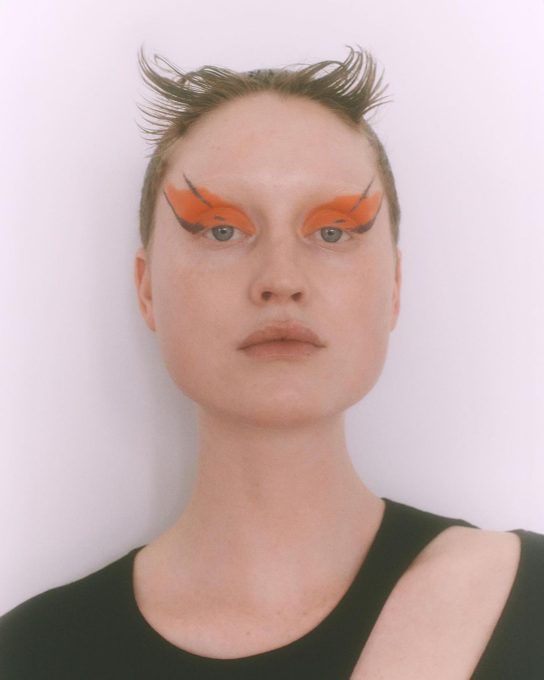 Close-up portrait of an adult woman with unique orange and black eyeshadow, short dark hair, and a black top against a white background.