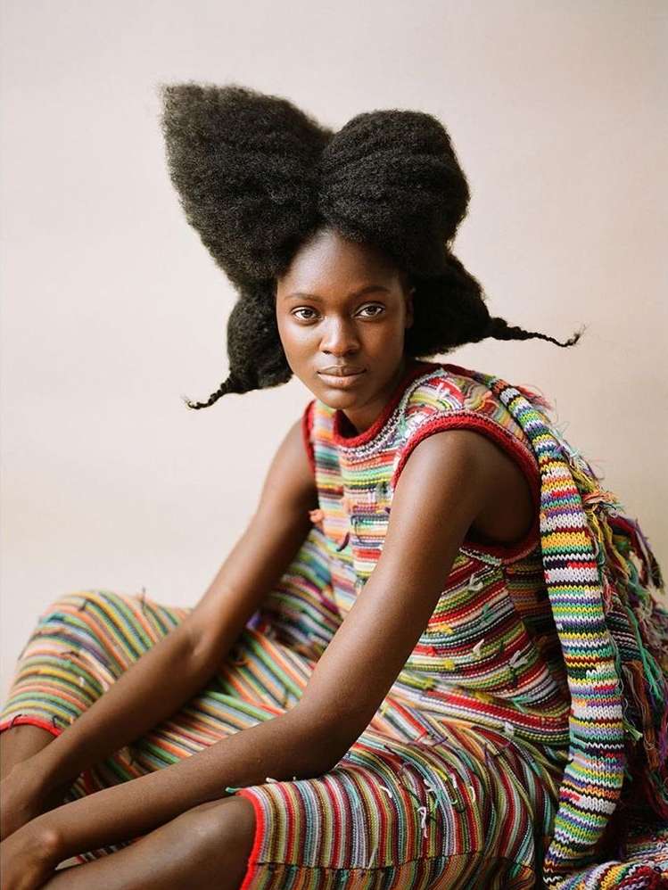 Portrait of an African woman with an elaborate afro hairstyle wearing a colorful striped dress in a neutral indoor setting.