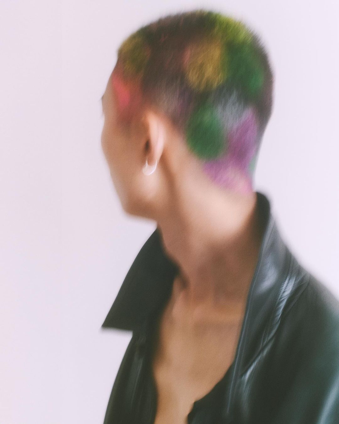 An adult male with a shaved head shows rainbow-colored spots, wearing a black jacket, with a silver earring, against a white background.
