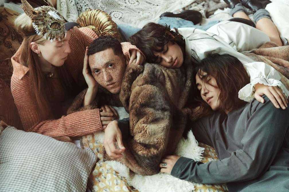 A cozy scene features a group of four people of diverse ethnicities relaxing together on a bed with blankets and pillows in a warm, inviting setting.