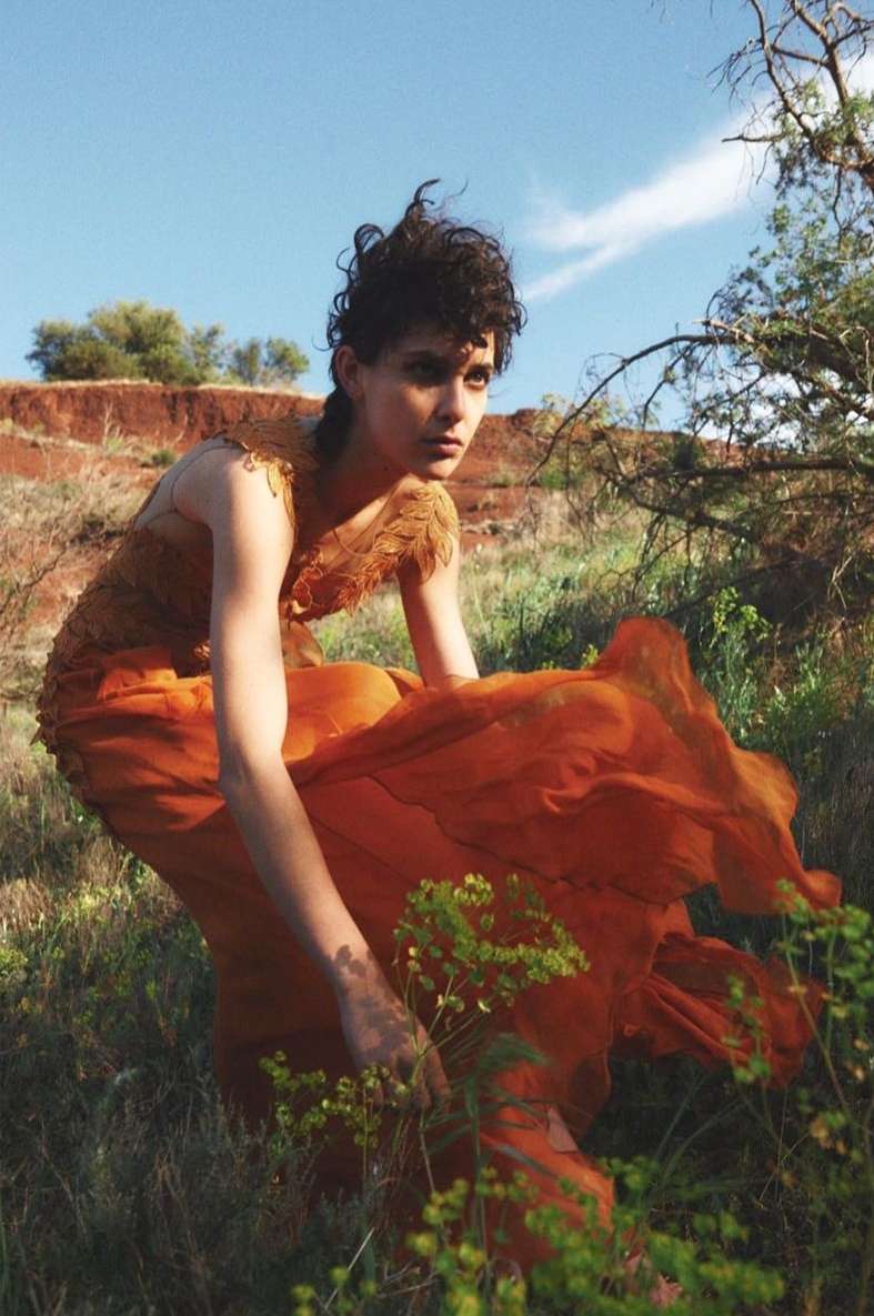 A young woman in an orange dress stands in a grassy field under a blue sky with light clouds. She is looking off to the side with a neutral expression.