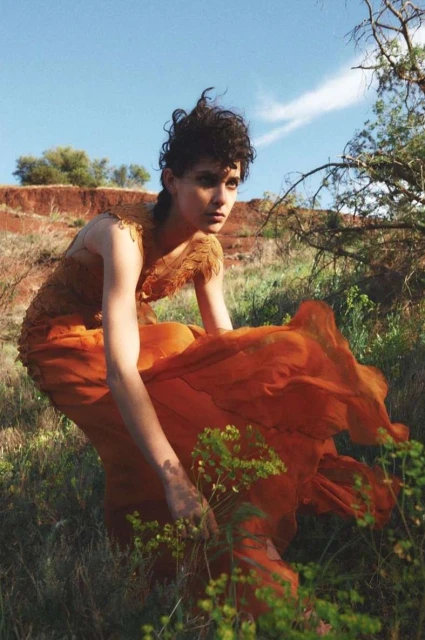 A young woman in an orange dress stands in a grassy field under a blue sky with light clouds. She is looking off to the side with a neutral expression.