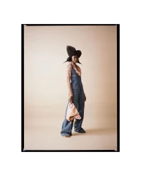 Model with sculptural black hair wearing a denim jumpsuit and patterned blouse against a plain grey studio backdrop