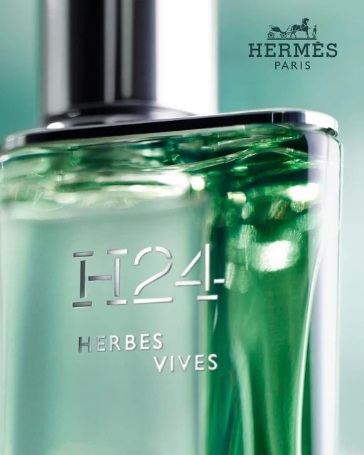 Close-up of a clear glass perfume bottle with a silver cap, featuring the Hermès logo and the words 'H24 Herbes Vives' in silver text.