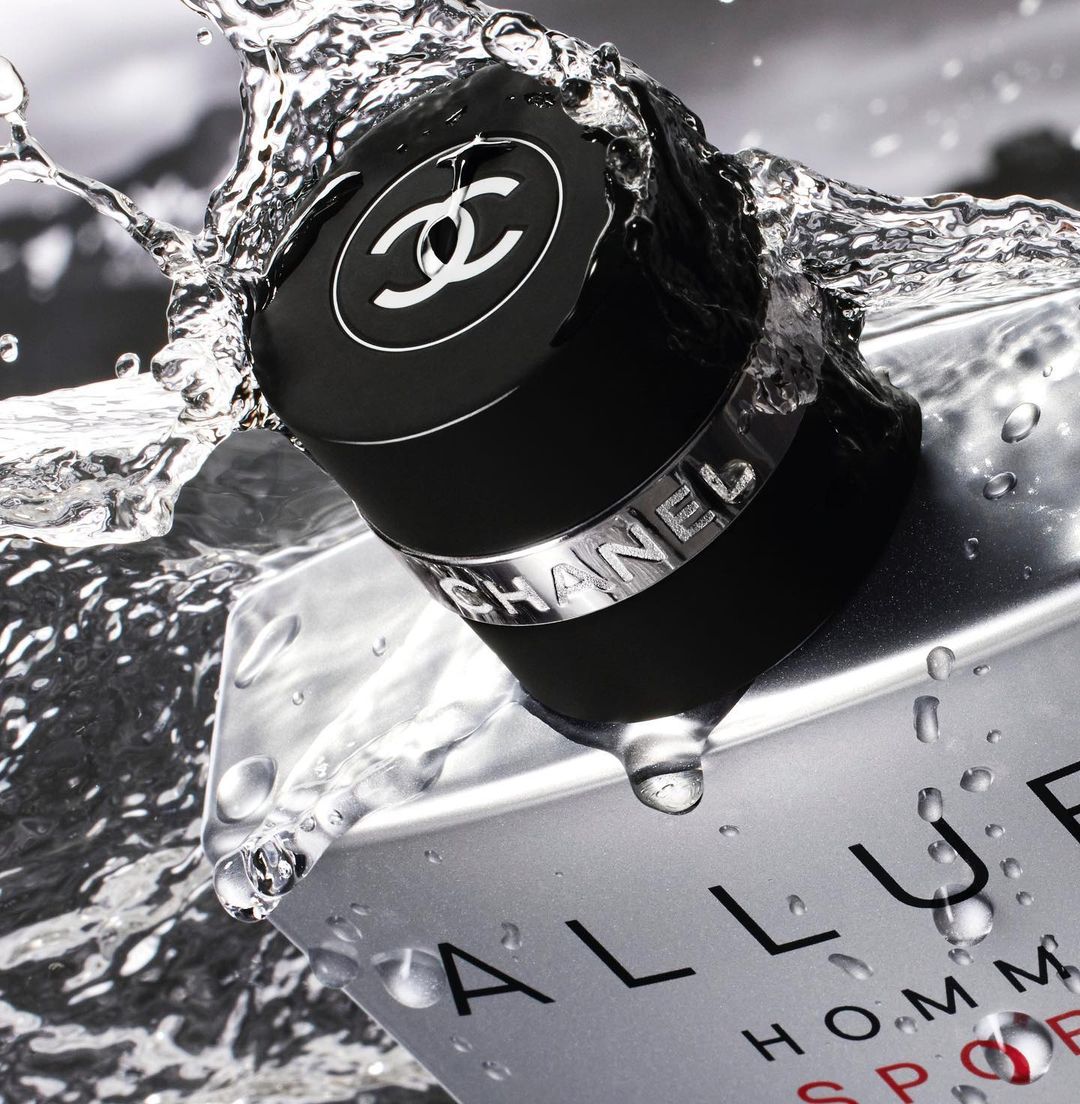 Close-up of a bottle of Chanel Allure Homme Sport perfume with water splashing around it, set against a blurred monochrome background.