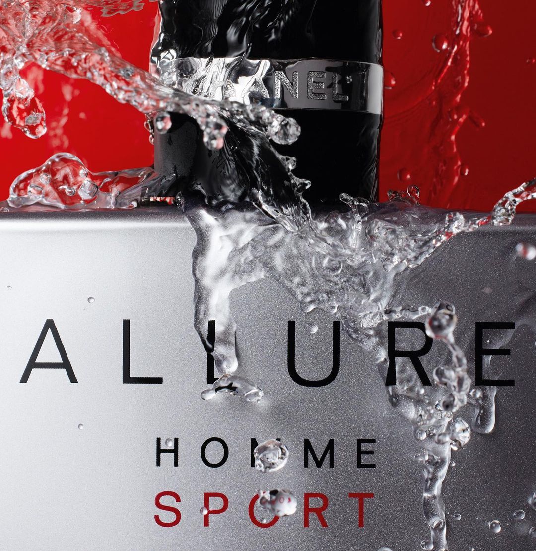 A bottle of Allure Homme Sport cologne is splashed with water against a gradient red and silver backdrop, featuring black typography.