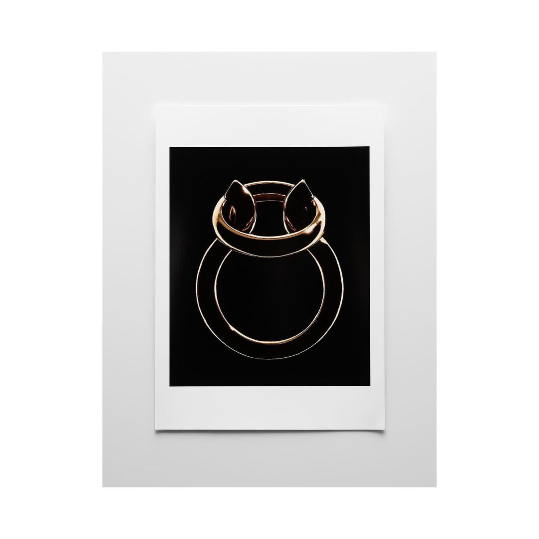 Abstract studio shot of a gold metal ring against a black backdrop, presented in a white frame. Simple, modern composition.
