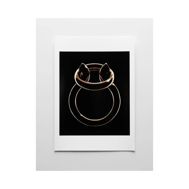 Abstract studio shot of a gold metal ring against a black backdrop, presented in a white frame. Simple, modern composition.