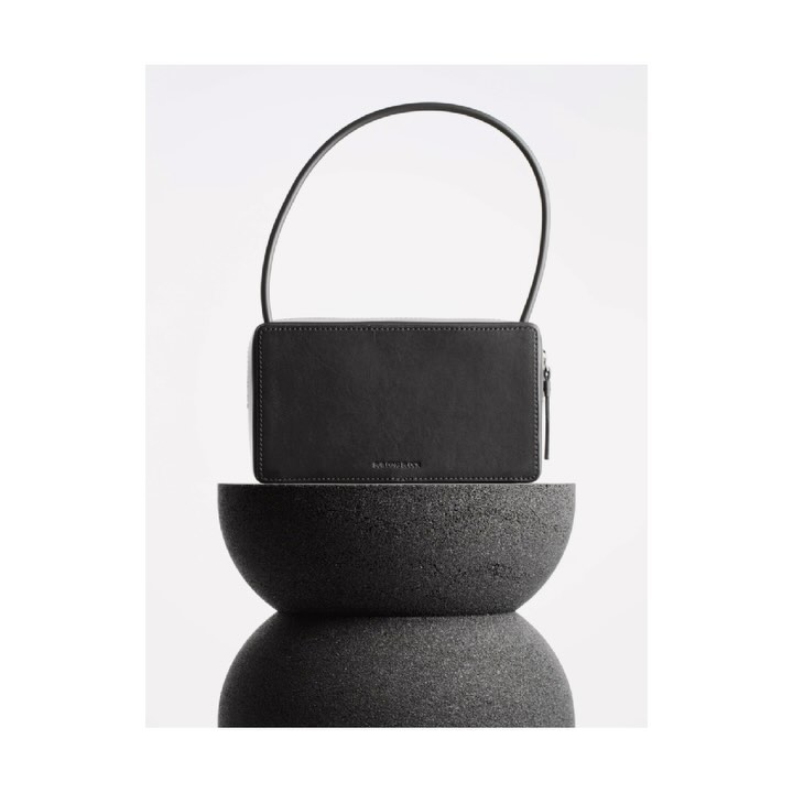 Studio shot of a black handbag with a handle on top of a stacked dark gray stone stand against a seamless bright white backdrop.