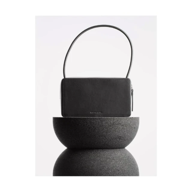Studio shot of a black handbag with a handle on top of a stacked dark gray stone stand against a seamless bright white backdrop.