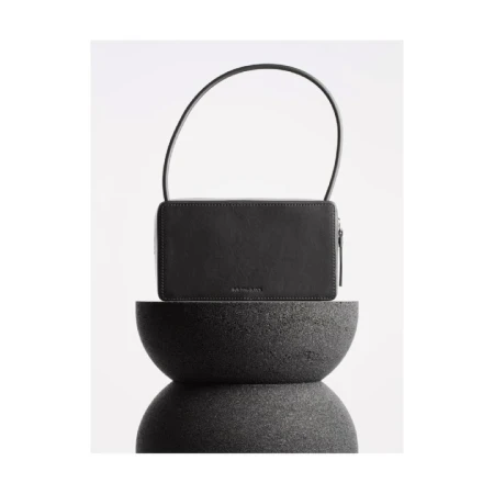 Studio shot of a black handbag with a handle on top of a stacked dark gray stone stand against a seamless bright white backdrop.