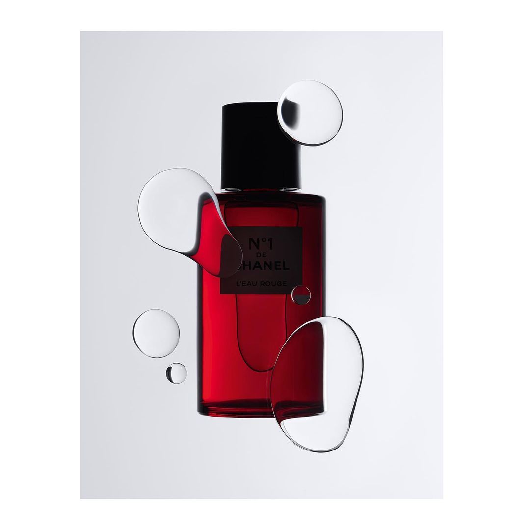 Close-up studio shot of a red glass Chanel No. 1 perfume bottle with black cap, surrounded by transparent liquid spheres, on a white background.