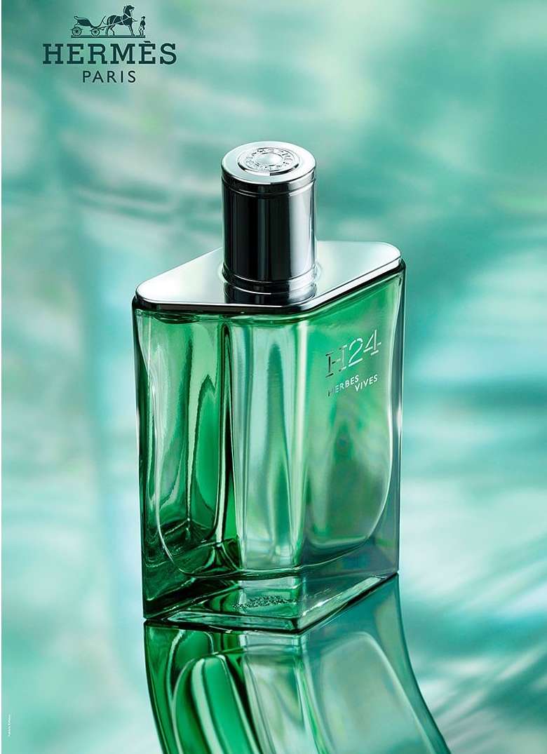 Green glass perfume bottle with silver cap on a reflective surface against a teal background, featuring the Hermes logo.