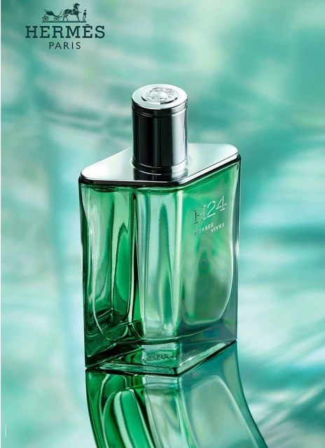 Green glass perfume bottle with silver cap on a reflective surface against a teal background, featuring the Hermes logo.