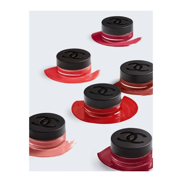 Overhead shot of six Chanel eye shadow containers with black lids and logo, arranged on a white surface with colorful smears of eyeshadow.