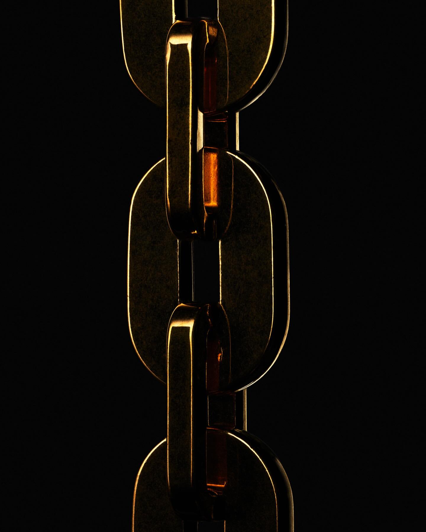 Macro shot of a gold metal chain link against a dark black background. The links catch light, showing their metallic texture and depth.