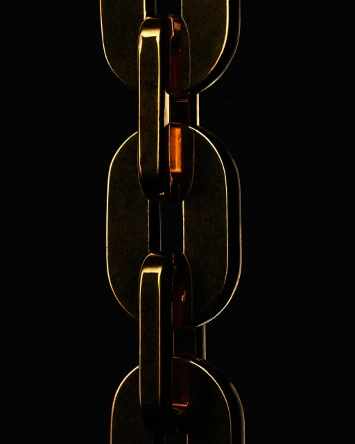 Macro shot of a gold metal chain link against a dark black background. The links catch light, showing their metallic texture and depth.