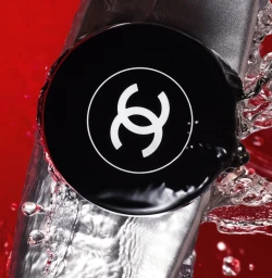 Close up of a black Chanel cosmetics compact surrounded by splashing water against a vibrant red background.