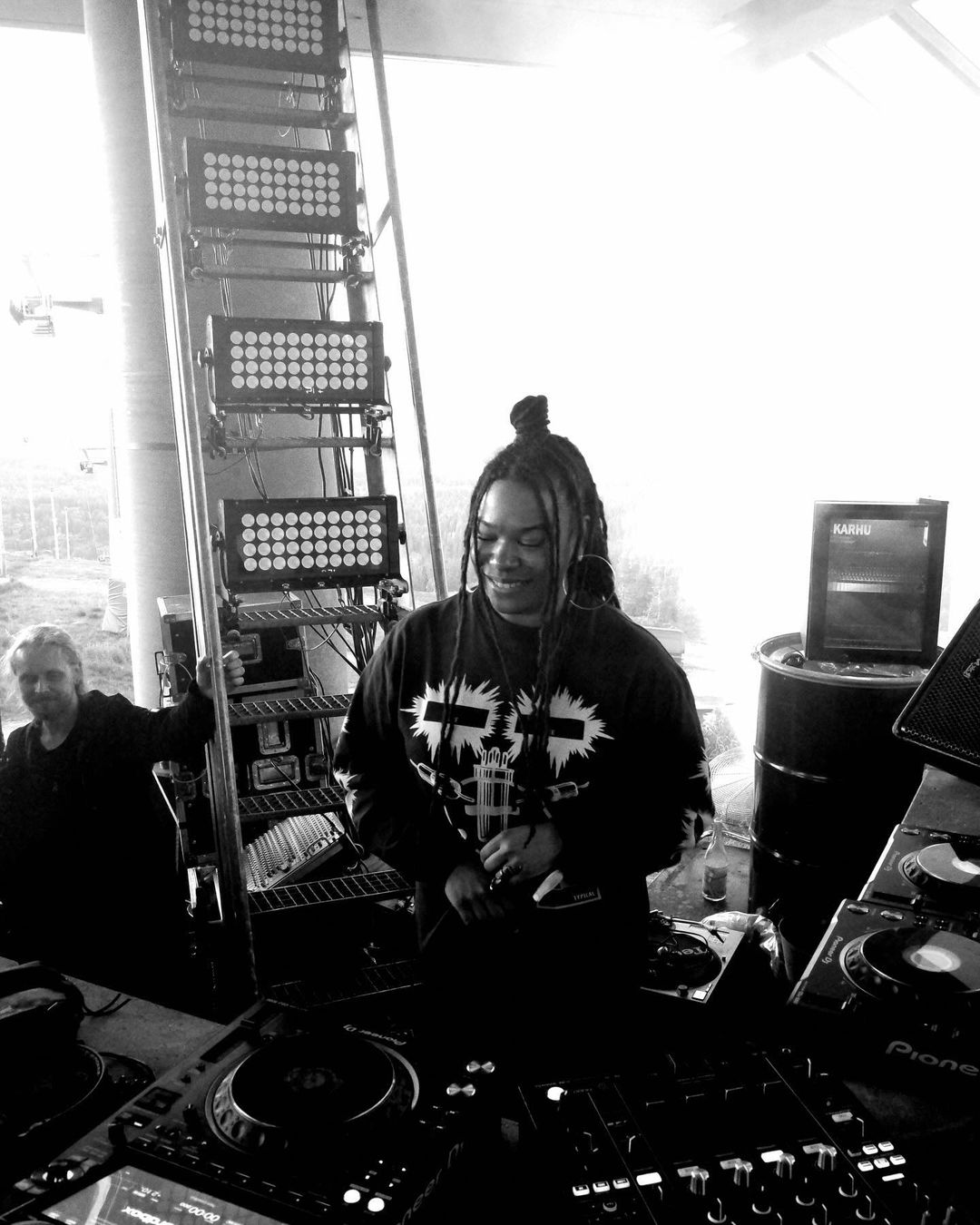 Black and white image of a female DJ working at her equipment with another person and lighting rig visible in the background.