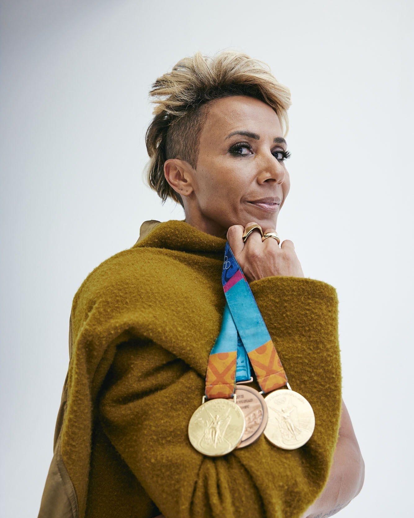 Portrait of an adult woman with an asymmetrical blonde hairstyle wearing a gold sweater and multiple Olympic medals, against a white background.