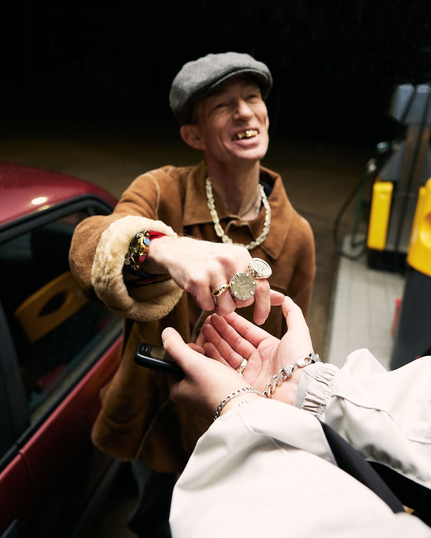 Two people with jewelry exchanging an object outdoors near a car and gas pump in dim light.