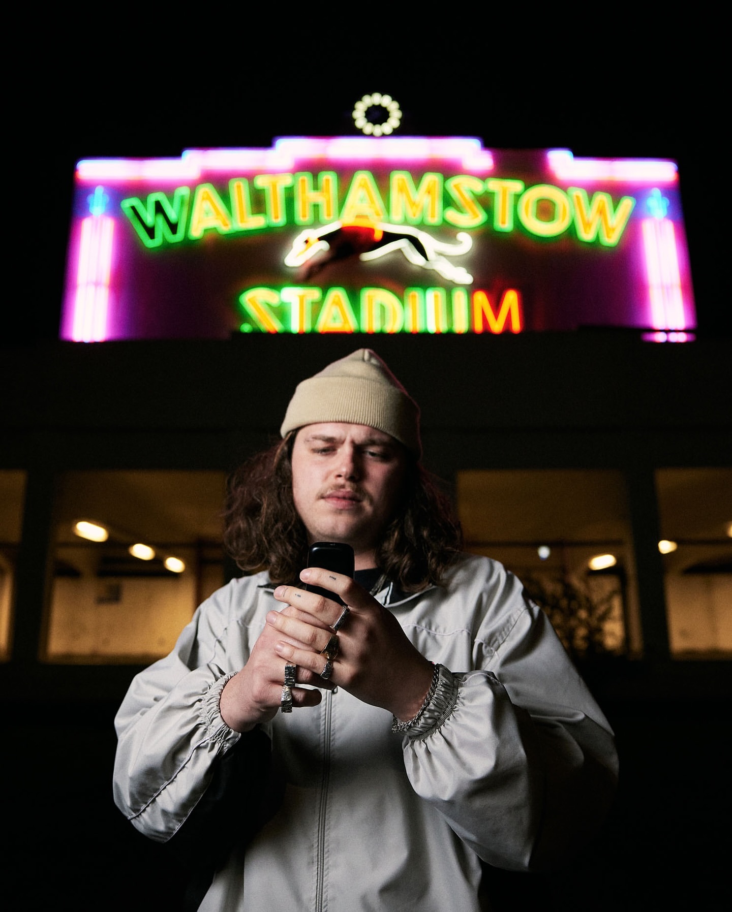 Man in a beanie using a phone at night in front of Walthamstow Stadium sign with neon lights, wearing a light jacket and rings.