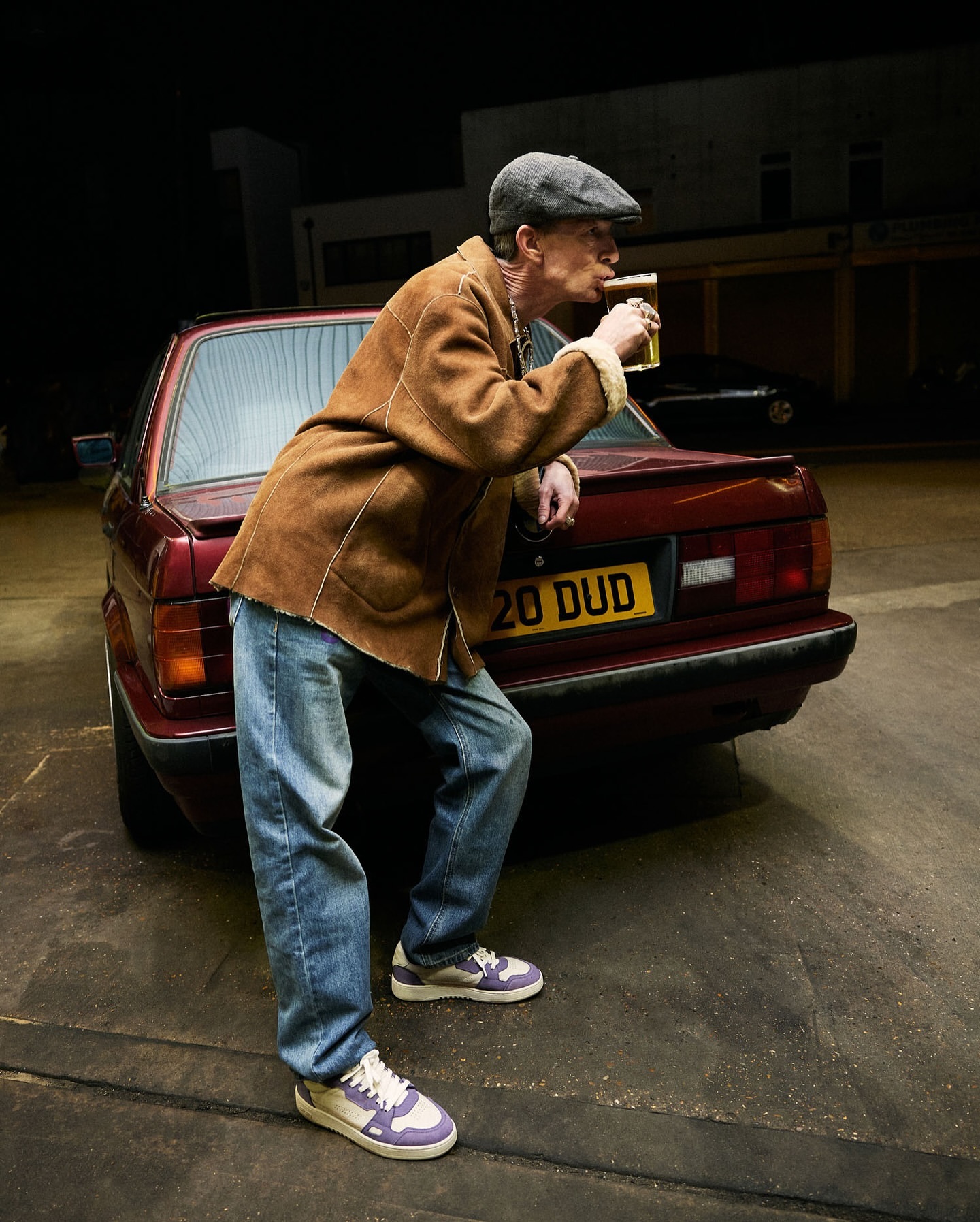 An older man in a cap and jacket leans against the back of a maroon car while drinking from a glass outdoors at night.