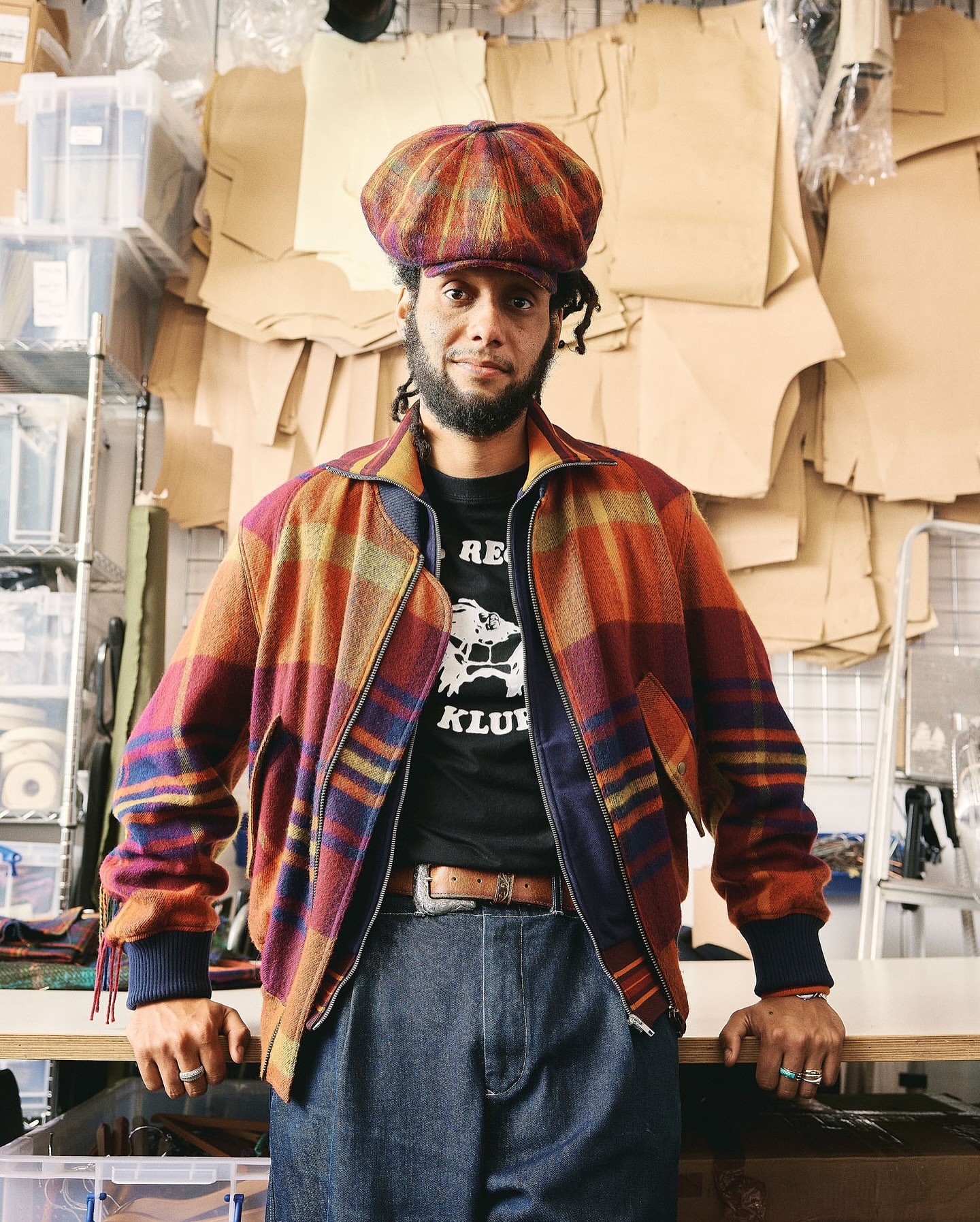 Adult male in a plaid jacket and cap poses indoors with a neutral expression against a backdrop of paper sewing patterns.