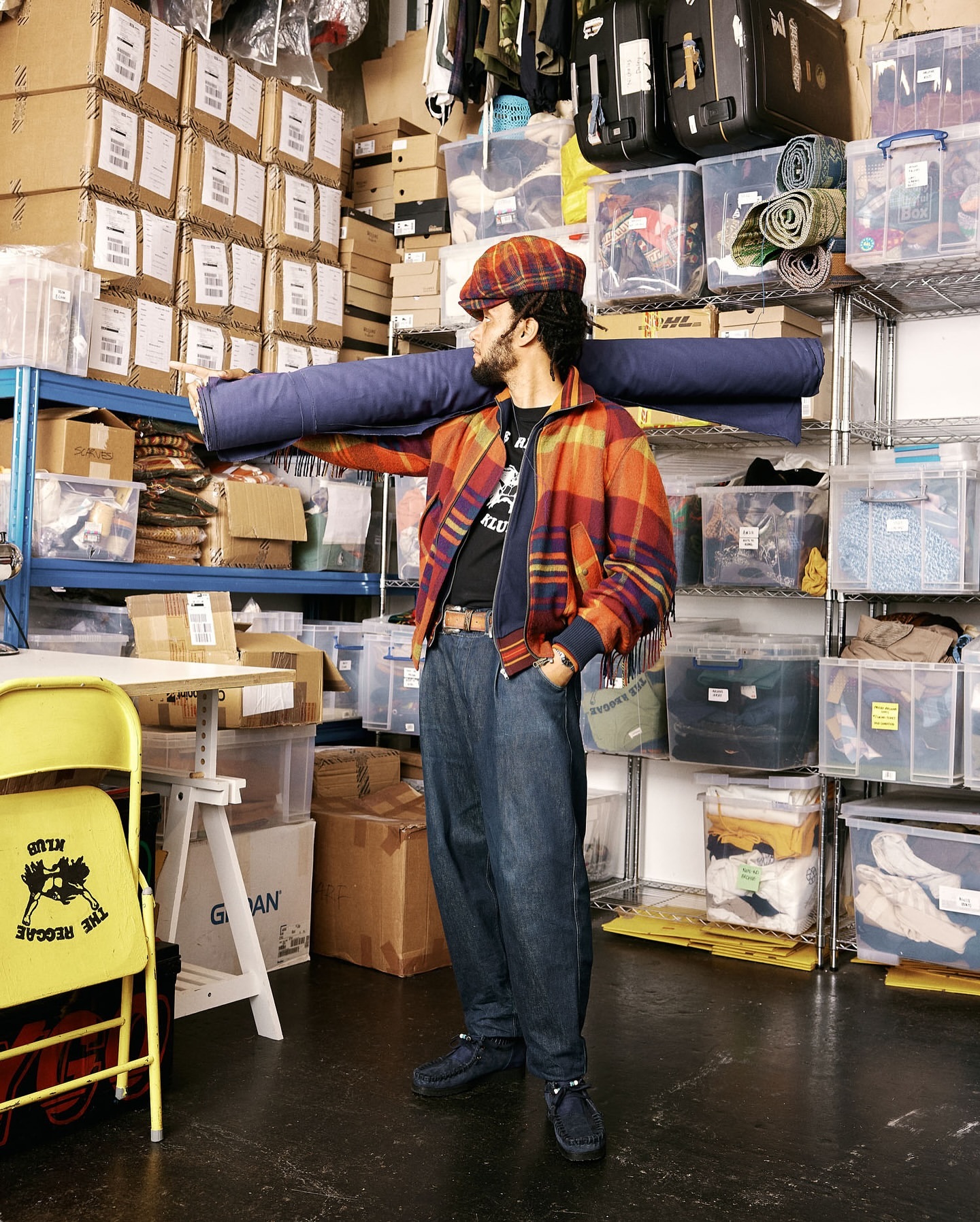 An adult man in a plaid jacket and hat holds a roll of blue fabric in a room filled with boxes and shelves of items in storage.