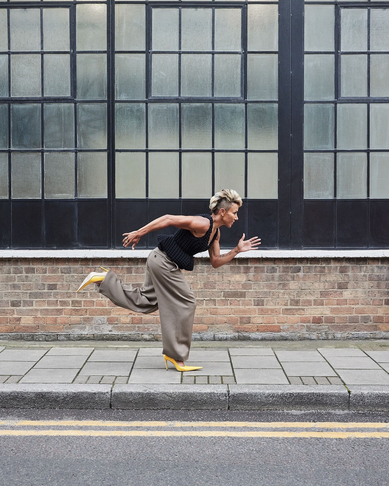 Adult woman with mohawk wearing a black top, tan pants and yellow heels runs along a city sidewalk with a large window in the background.