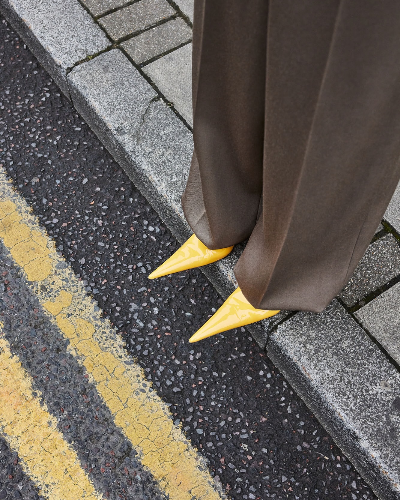 A person stands on a city street wearing brown trousers and yellow pointed heels, with grey pavement and yellow road markings.