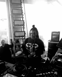 Black woman DJ with braided hair wearing a dark graphic sweatshirt standing behind mixing decks in London