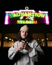 Man wearing beanie and light jacket looking down at mobile phone beneath glowing Walthamstow Stadium neon sign