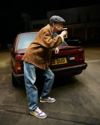 An older man wearing a flat cap, brown shearling jacket, and blue jeans leans against the trunk of a red car while drinking from a pint glass.