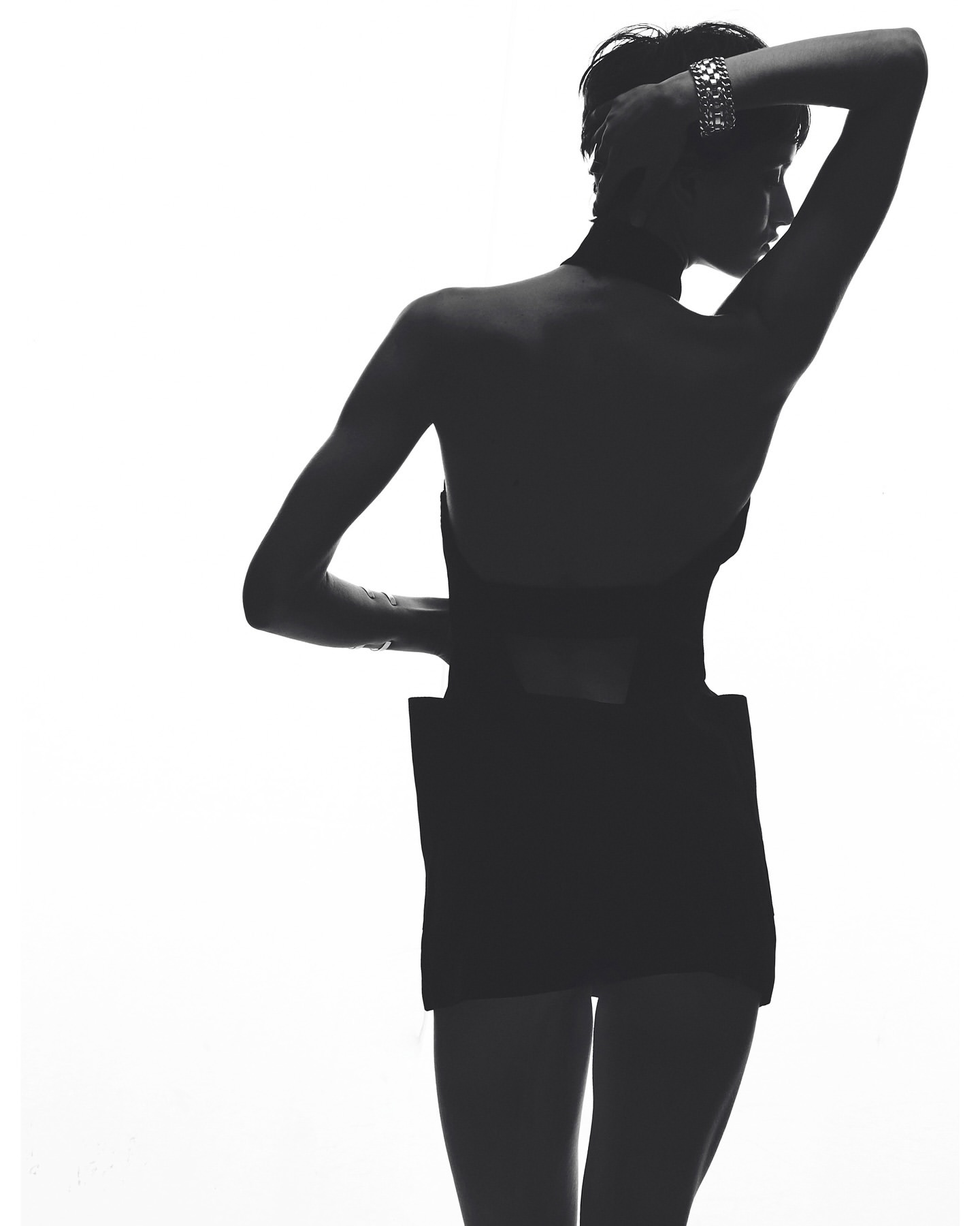 Backlit studio shot of a silhouette of a woman with short hair, wearing a black dress and silver bracelet against a white background.