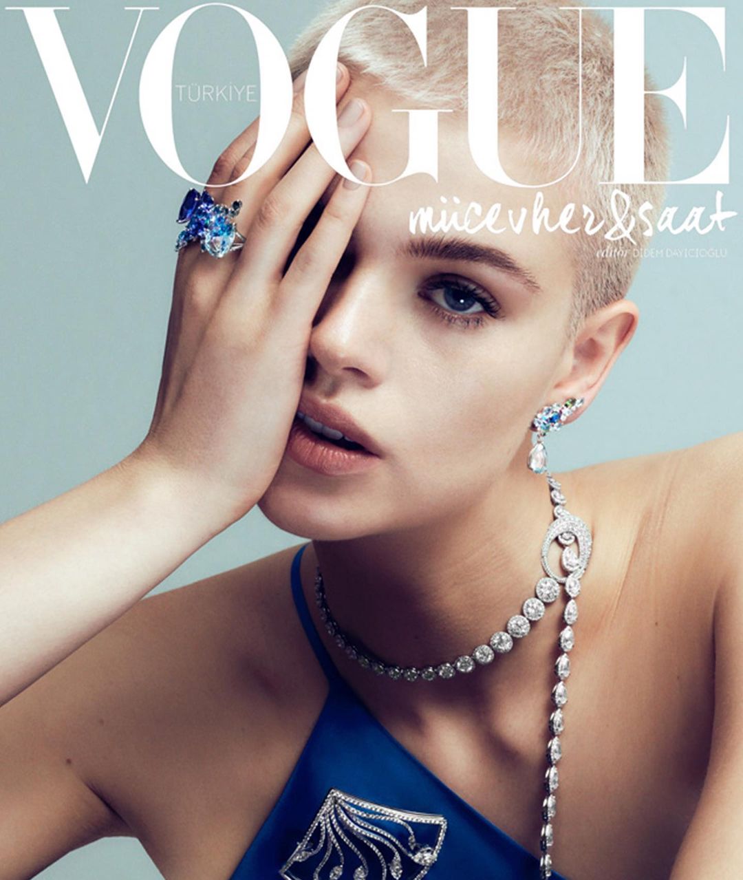 Close-up portrait of a blonde model wearing a blue top, ornate diamond necklace, and large gemstone ring, photographed by Onur Dag.