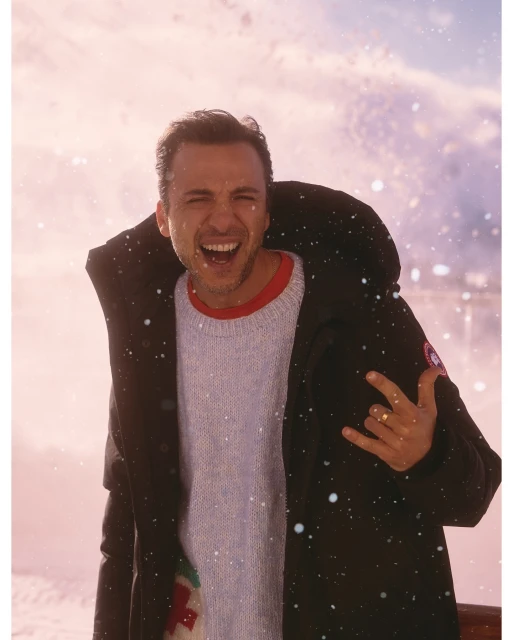 A man smiles in a black winter coat, light blue sweater, and red undershirt with a blurry snow-covered background in a medium shot.